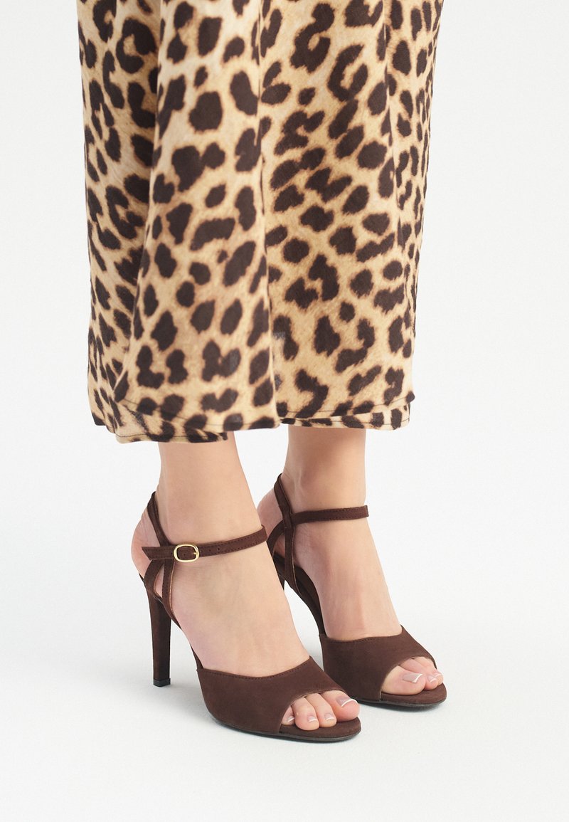 Sandalias de tacón alto de ante marrón con diseño de punta descubierta, tira en el tobillo y detalle de hebilla dorada. El fondo presenta una tela de estampado de leopardo.
