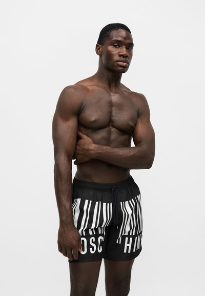 Homme musclé torse nu portant un short rayé noir et blanc avec un cordon, debout devant un fond clair uni.
