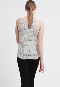 Top senza maniche grigio chiaro in maglia con un motivo a strisce testurizzato. Materiale morbido con una trama aperta, caratterizzato da un orlo arrotondato. Indossato con pantaloni neri.