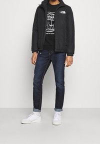 Czarna pikowana kurtka na czarnym T-shircie z grafiką, w połączeniu z ciemnymi jeansami i białymi trampkami. Prosty fason i luźny krój.