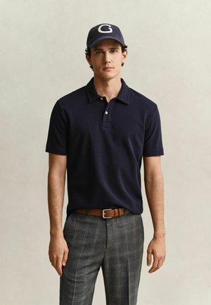 Mann trägt ein marineblaues Polohemd, eine marineblaue Kappe mit "G,", einen braunen Gürtel und graue karierte Hosen und steht vor einem einfarbigen Hintergrund.