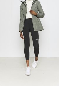 Grüne wasserdichte Jacke mit Kapuze, langärmliges weißes Oberteil, schwarze Leggings mit Logo und weiße Sportschuhe.