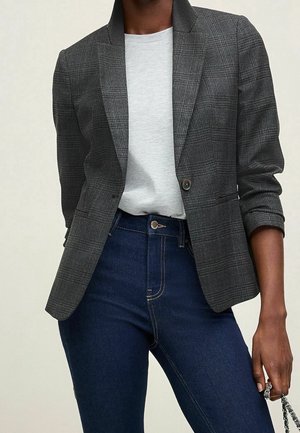 Osoba nosząca ciemnoszary kraciasty blezer na jasnoszarej koszuli i ciemnoniebieskich dżinsach z wysokim stanem, trzymająca w jednej ręce srebrny łańcuszek.