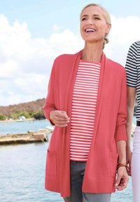 Gilet corail avec un col châle et des poches avant, superposé sur un haut rayé rouge et blanc. L'ensemble est associé à un pantalon gris.