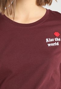 Camiseta burdeos con cuello redondo que presenta texto bordado en blanco "Kiss the world" y un gráfico de labios rojos. Tela de algodón suave.