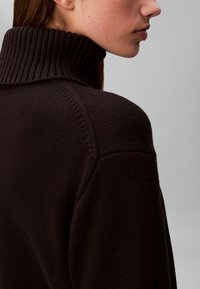 Woman wearing a dark brown knitted turtleneck sweater shown from the side focusing on the shoulder and collar area.