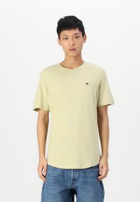 Svijetložuti t-shirt kratkih rukava s okruglim izrezom i malim crnim logom na prsima, u kombinaciji s plavim trapericama širokog kroja.