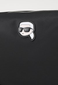 Černý zipový sáček s stylizovanou tváří s bílými vlasy, která má na sobě velké černé sluneční brýle, umístěnou ve středu přední strany.