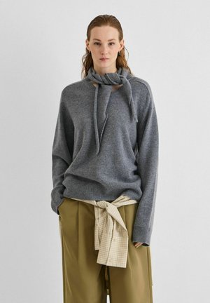Mujer lleva un suéter de punto gris con detalle en el cuello y pantalones sueltos verde oliva con una camisa beige de cuadros atada alrededor de la cintura.