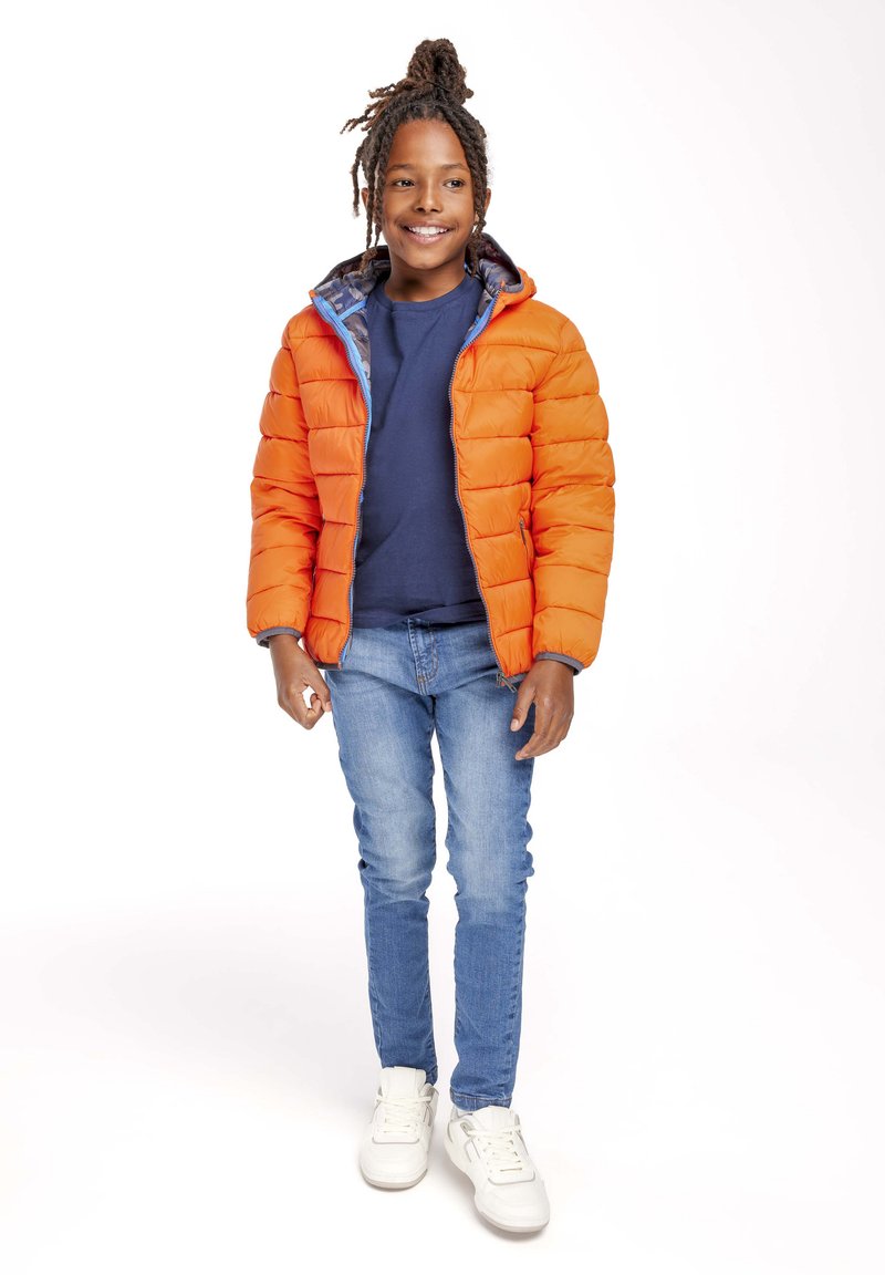 Giacca a vento arancione con fodera interna blu, indossata sopra una camicia blu navy, abbinata a jeans blu e sneakers bianche.