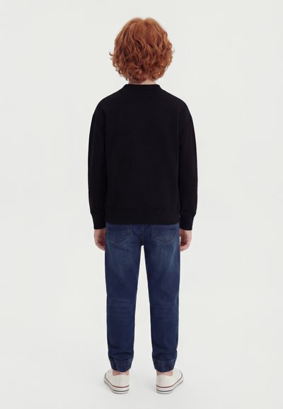 Παιδί με σγουρά κόκκινα μαλλιά στέκεται γυρισμένο με την πλάτη, φορώντας μαύρο φούτερ, μπλε τζιν με ελαστικές μανσέτες και λευκά αθλητικά παπούτσια.