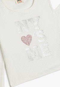 Weißes geripptes Oberteil mit großen silbernen Paillettenbuchstaben, die "NY'S ME" buchstabieren, mit einem roten Herzdetail in der Mitte.