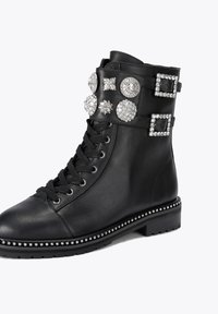 Stivaletto in pelle nera presenta una punta arrotondata, design con lacci, cinghie decorate con cristalli e borchie decorative lungo la suola.