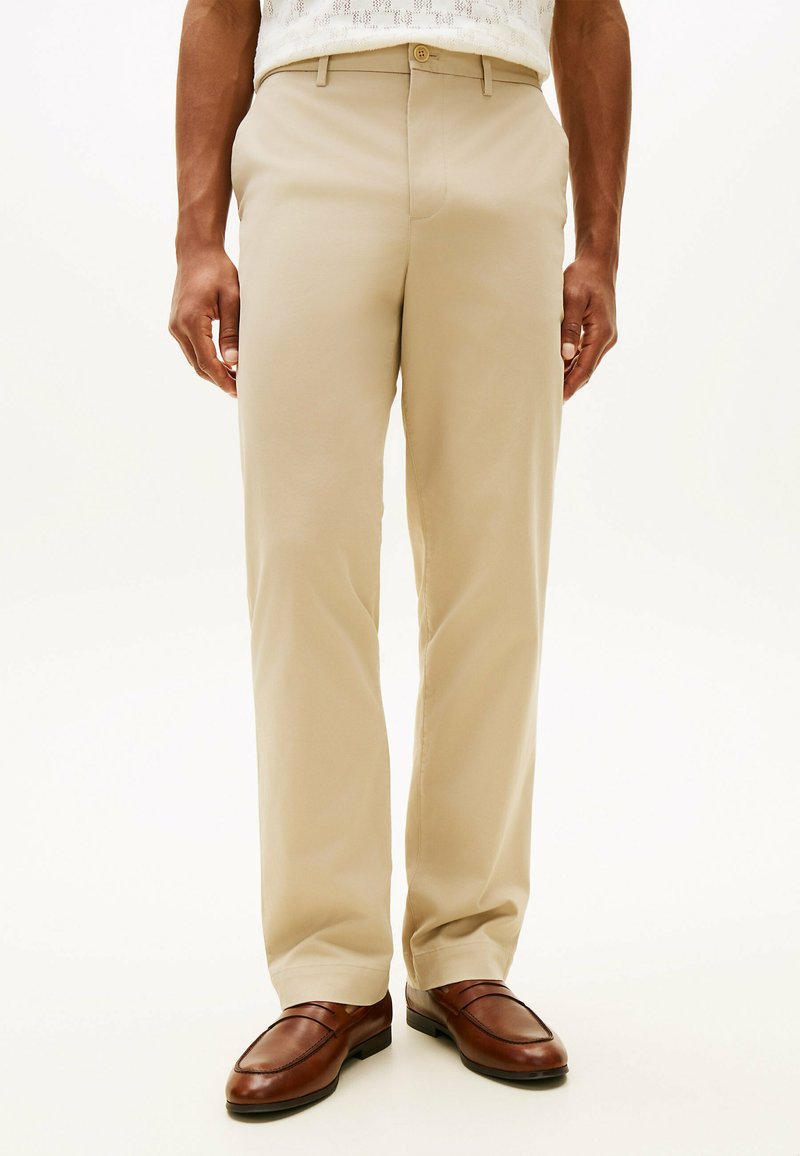 Uomo che indossa pantaloni beige e mocassini di pelle marrone, in piedi con le mani rilassate lungo i fianchi su uno sfondo bianco.