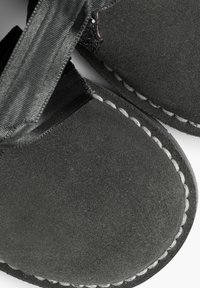 Zapatillas de ante negro con puntera redondeada y costuras blancas visibles a lo largo de los bordes. Presentan una textura suave y una cinta de tela.