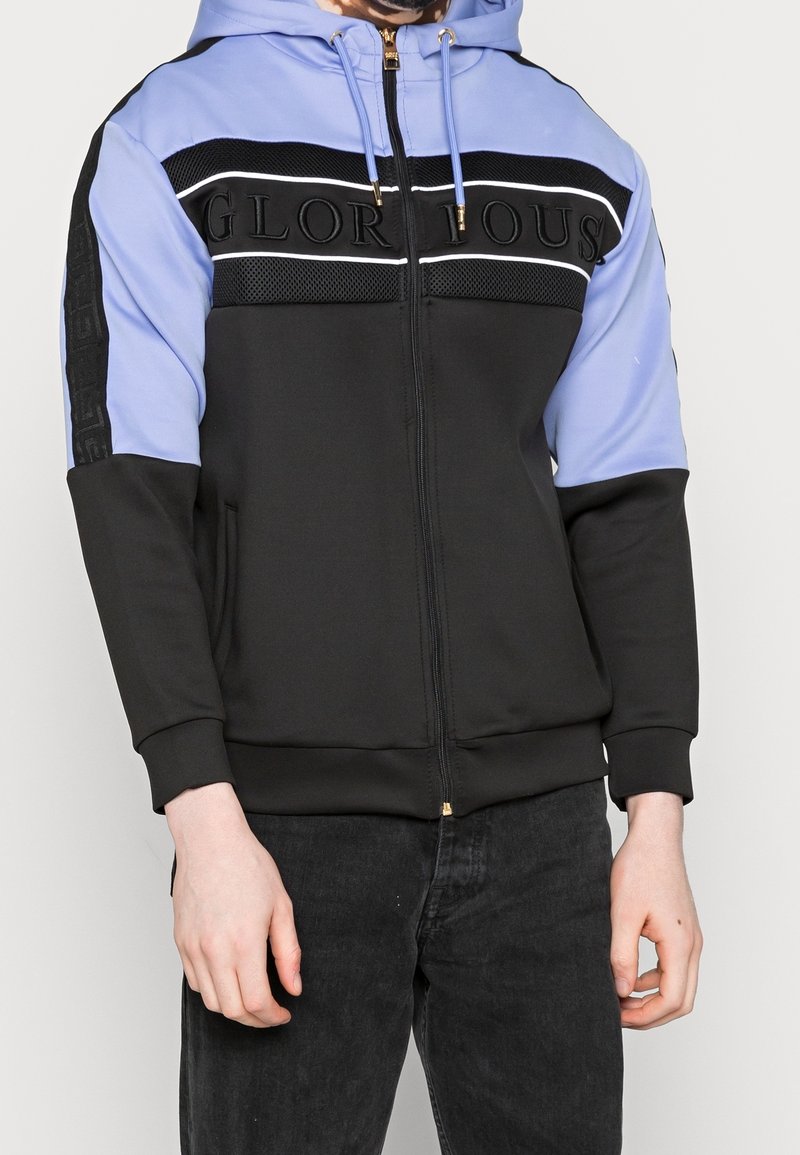 Homme portant un sweat à capuche zippé noir et bleu clair avec le texte "GLORIOUS" sur la poitrine et un jean noir, debout devant un fond uni.