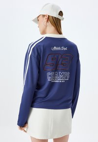 Tummansininen urheilullinen pitkähiha, jossa on valkoiset raidat, ja jossa on rohkeasti painettu "93 CHAMPS" sekä "Athletic Dept" -logo takana. Sujuva pinta.