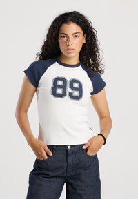 Camiseta blanca ajustada de manga corta con mangas raglán en color azul marino y gráfico frontal del número "89" en un azul marino contrastante. Lleva unos jeans de rayas azules.