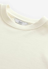 Cremefarvet bomulds T-shirt, afslappet pasform, rund halsudskæring, blød tekstur, subtile syninger og et mærke med "NEXT" branding synligt.