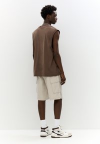 Maglietta senza maniche marrone, pantaloni cargo beige con tasche laterali, calzini bianchi e sneakers, visti da dietro su uno sfondo chiaro.