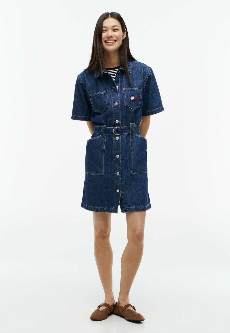 Femme debout les mains dans le dos, portant une robe en jean ceinturée à manches courtes avec des boutons et des chaussures marron à enfiler, souriante.