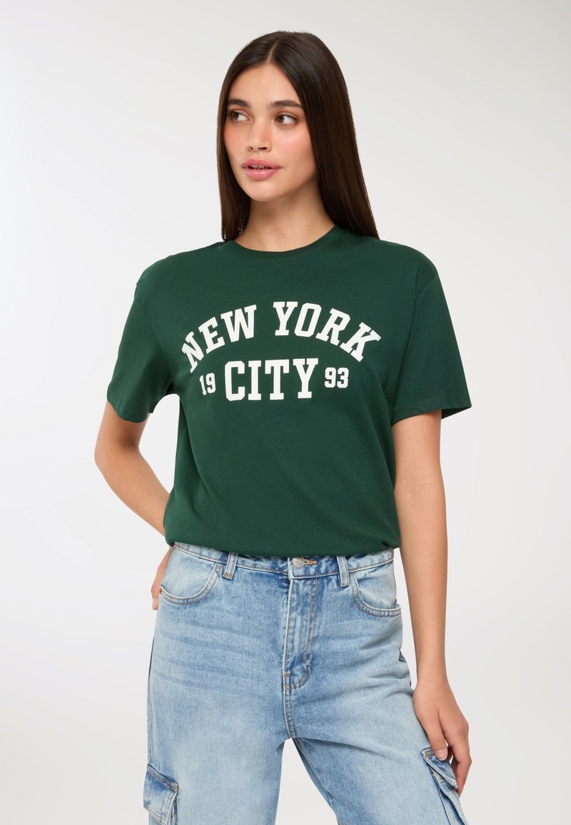 Giovane donna con lunghi capelli scuri che indossa una maglietta verde "New York City 1993" e jeans cargo blu chiaro, mentre guarda a sinistra.