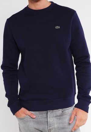 Sudadera azul marino hecha de tela suave, con cuello redondo y puños acanalados. Incluye un pequeño logo de cocodrilo verde en el pecho.