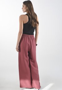 Wijdvallende broek van gestructureerde roestrode stof met een elastische tailleband, gecombineerd met een aansluitend zwart topje, gezien van achteren.