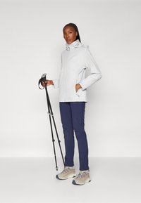 Ljusgrå vattentät jacka med huva, kombinerad med marinblå byxor och beige vandringskängor. Modellen håller i svarta vandringsstavar.