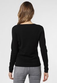 Schwarzer Langarmpullover mit Rundhalsausschnitt, mit einer figurbetonten Passform und gerippten Bündchen. Trägt dazu eine hellgraue, enganliegende Jeans.