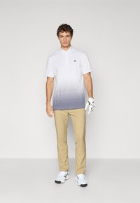Muž v bílém polo tričku s gradientem, béžových kalhotách, bílých golfových rukavicích a bílých teniskách, stojící na jednobarevném světlém pozadí.