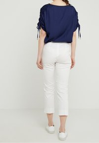 Woman standing facing away wearing a navy ruched short-sleeve blouse, white cropped pants, and white slip-on sneakers.