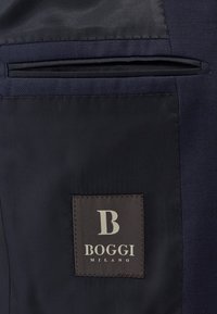 Marine stof met een gladde, matte textuur heeft een verticale zak en een bruine label met "BOGGI MILANO" in crèmekleurige letters.