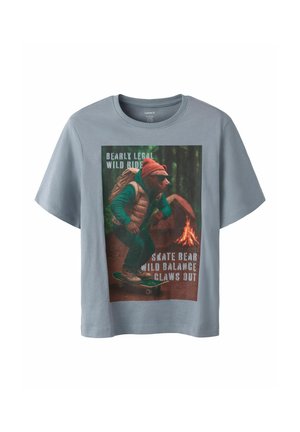 T-shirt bleu clair avec un imprimé d'un ours portant des lunettes de soleil, un sac à dos et un bonnet, faisant du skateboard dans une forêt près d'un feu de camp.