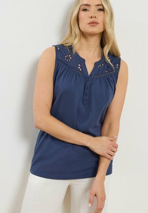 Donna bionda con top blu navy senza maniche, con intagli floreali vicino alle spalle e scollo a V con bottoni, abbinato a pantaloni bianchi.