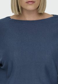 Bleden modra, teksturirana pletena majica s širokim izrezom in ohlapnim krojem. Mehka tkanina z rahlim raztezanjem, brez vidnih vzorcev ali okraskov.