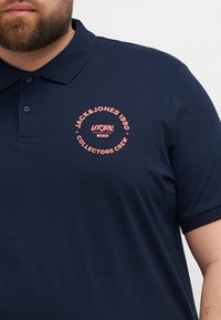 Tricou polo bleumarin din amestec de bumbac, cu un logo circular roz cu text pe pieptul stâng, guler cu două butoane și mâneci scurte.