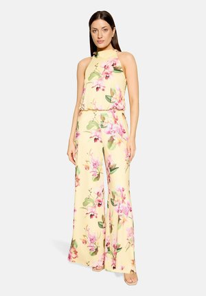 Kobieta ubrana w żółty, bezrękawnikowy kombinezon z różowo-zielonym kwiatowym wzorem, stoi z rękami swobodnie opuszczonymi wzdłuż ciała.