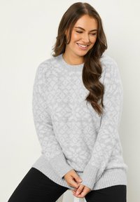 Jasnoszary sweter z teksturowanym wzorem, z prążkowanymi mankietami i dołem, oraz okrągłym dekoltem, noszony na czarnych spodniach.