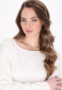 Bílý dlouhý rukáv top s širokým výstřihem a elastickými manžetami, hladká tkanina a jemný splývavý střih. Modelka má dlouhé, vlnité hnědé vlasy.