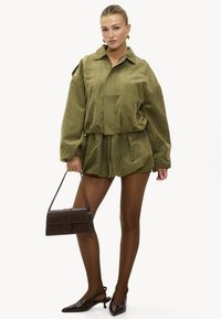 Ensemble chemise oversize et shorts vert olive en tissu doux, assorti à un sac à main marron et des chaussures à talons aiguilles pointues. Collants transparents.