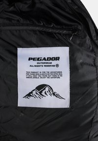 Svart nylonjakkeinnside med en hvit etikett med svart tekst og fjell-logo, som gir produktinformasjon og pleieinstruksjoner.