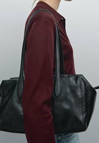 Zwarte leren handtas met twee lange hengsels, rechthoekige vorm, gladde textuur en gestikte details. Draagt over een bordeauxkleurige top.