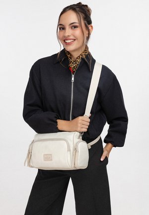 Jeune femme souriante portant une veste noire à fermeture éclair et une écharpe à imprimé léopard, tenant un sac bandoulière beige clair avec plusieurs poches zippées.