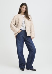 Veste matelassée beige à coupe oversize, col rond et fermeture par boutons-pression ; associée à un jean denim bleu foncé à jambes larges.