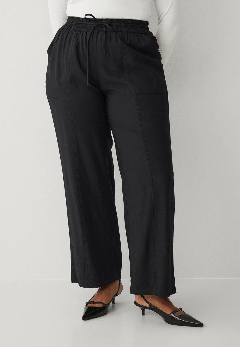 Pantalons larges noirs avec une taille élastique, un cordon de serrage et des poches latérales, présentant une texture lisse et un design minimaliste.