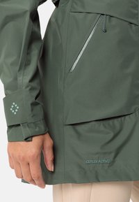 Giacca impermeabile verde con finitura morbida, dotata di tasca con zip, polsini regolabili e marchio Ceplex Active in azzurro chiaro.