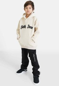 Felpa beige con la scritta "Sixth June" in nero, abbinata a pantaloni cargo neri con cerniere e tasche, indossata con sneakers nere.