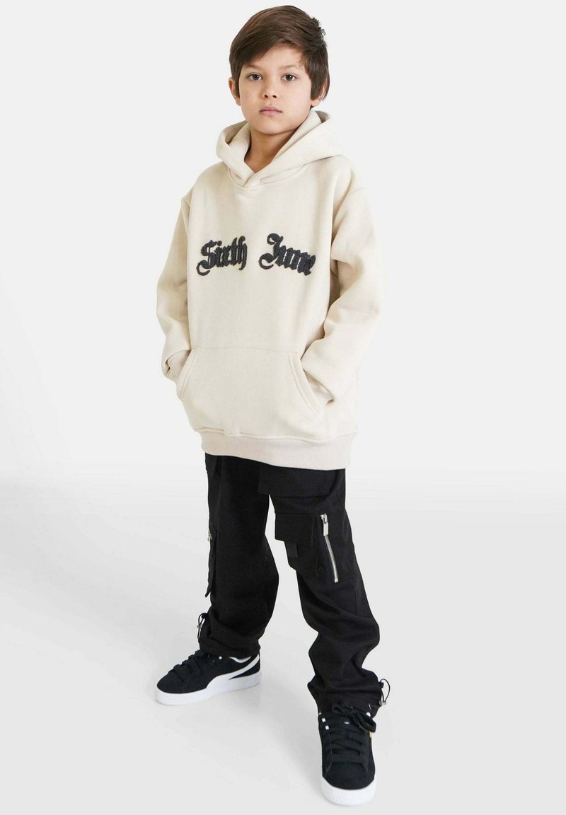 Felpa beige con la scritta "Sixth June" in nero, abbinata a pantaloni cargo neri con cerniere e tasche, indossata con sneakers nere.