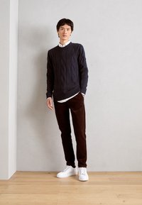 Marinblå kabelstickad tröja över en vit skjorta, kombinerad med bruna kordflorsbyxor och vita sneakers. Minimalistisk stil mot en neutral bakgrund.